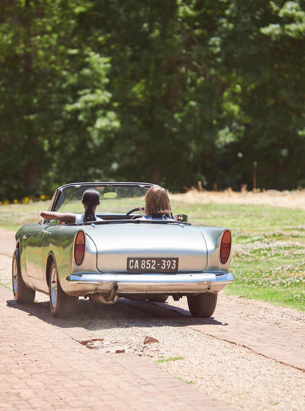 Two women drive away ina vintage car on a sunny day.