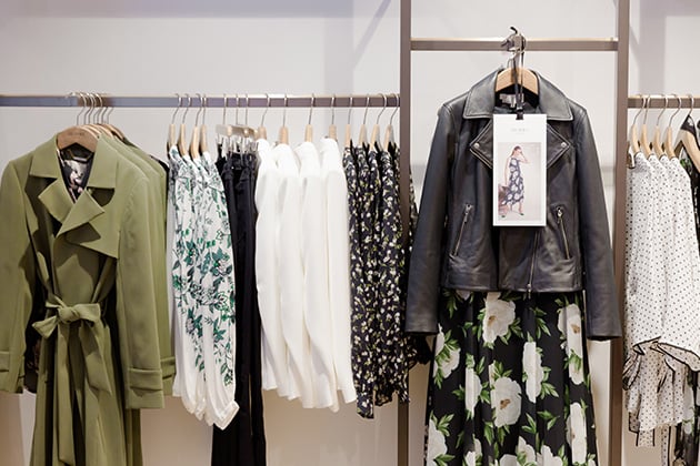 A clothing rail in a Hobbs store. The hanger in the middle shows a dogtooth patterned shirt dress in black and white with a crossbody leather bag in white. The hangers on either sides feature garments in black, white, grey and red.