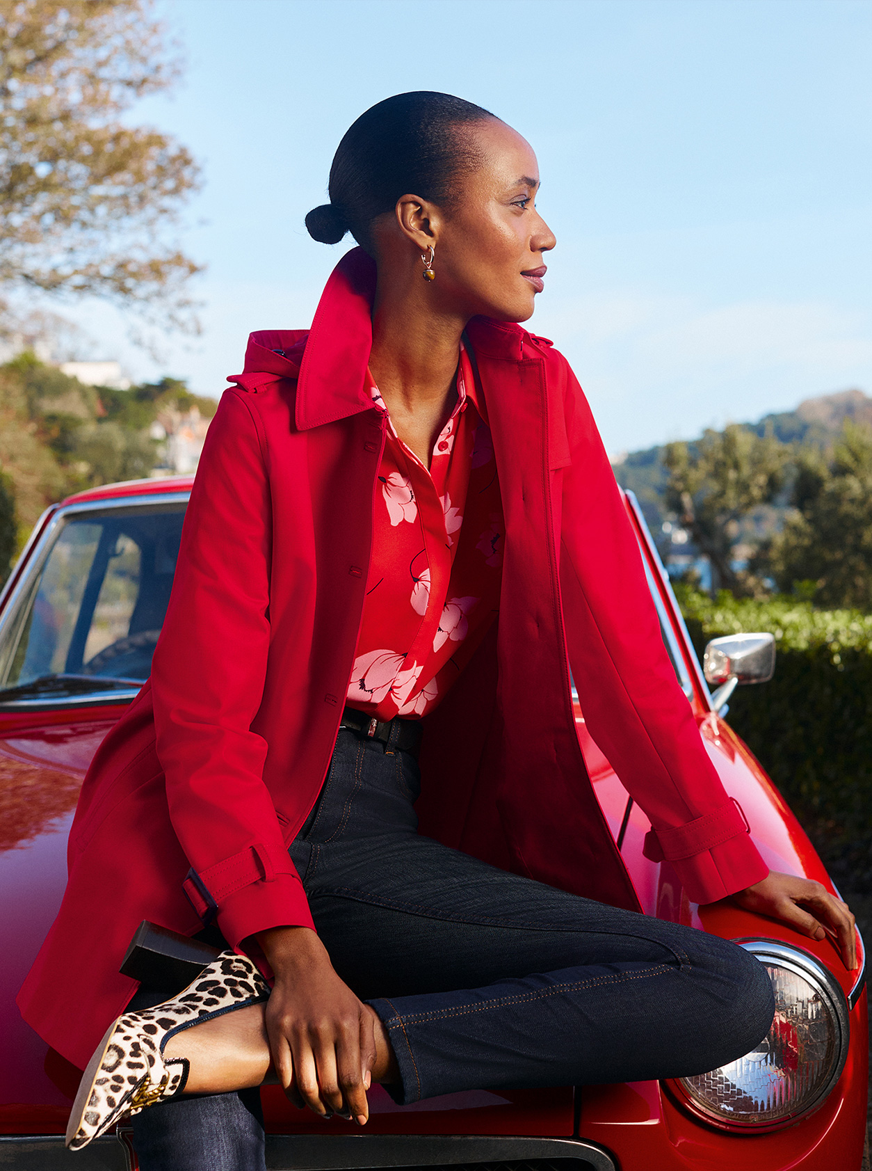 Model photographed sitting on the bonnet of a red car looking off to the right, wearing a Hobbs red trench coat, floral blouse and dark wash blue jeans.