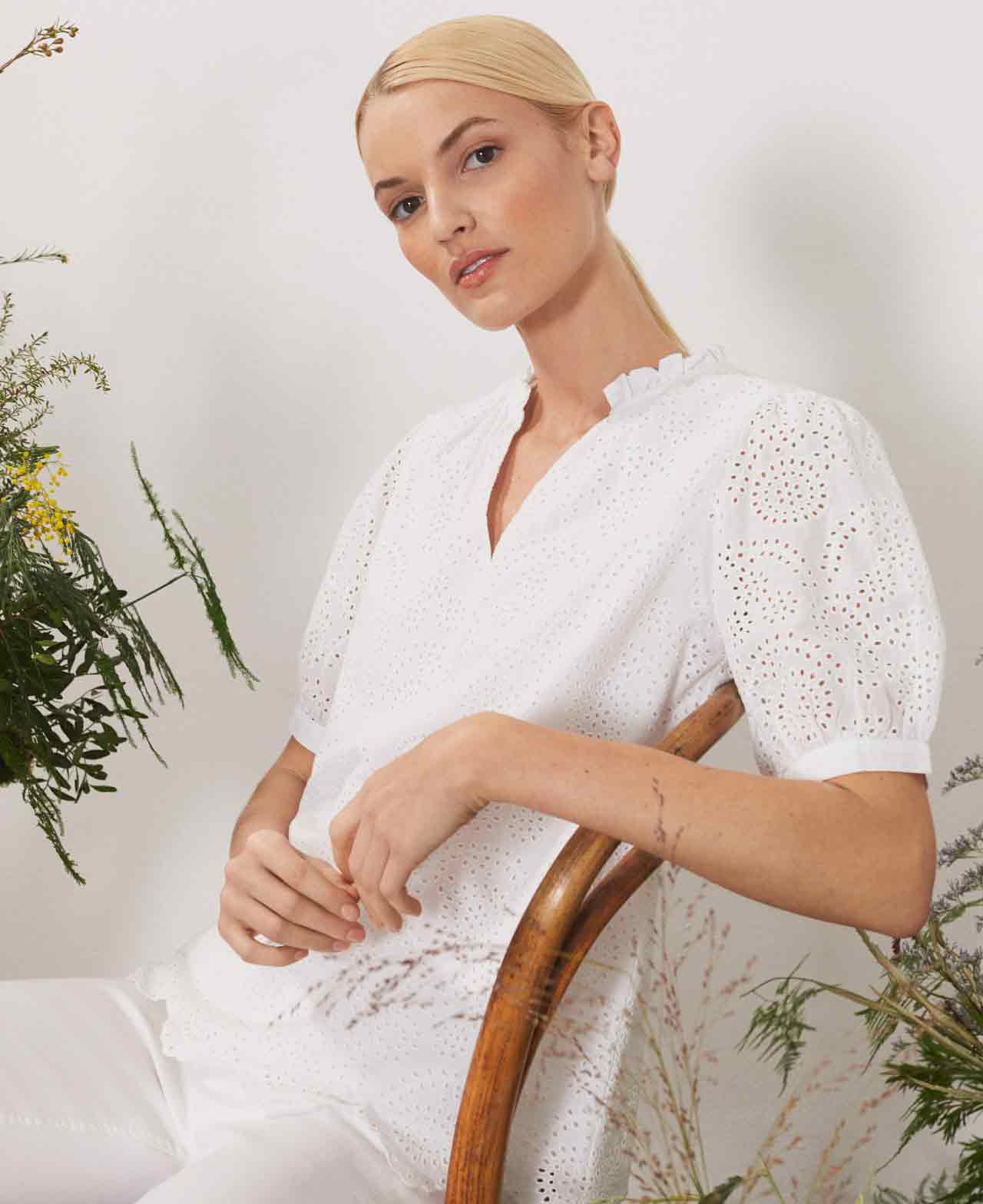 Hobbs model sits on a chair wearing a white broderie anglaise blouse.