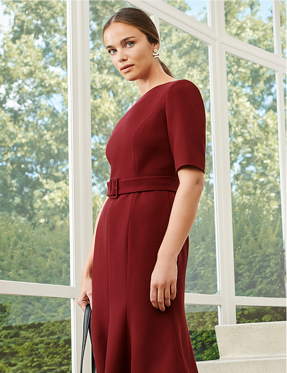 Model wears a bright red belted dress with a black patent leather bag