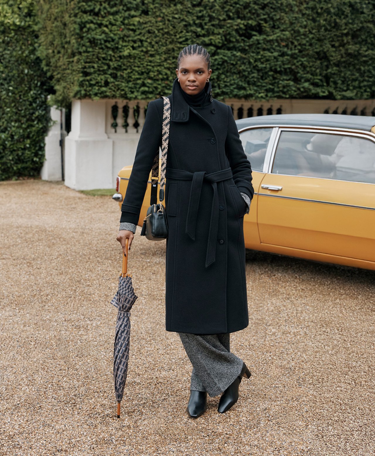  A woman wearing a long, black Hobbs coat, with a monogram umbrella and matching bag.
