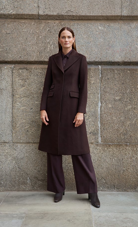 Model stands against a wall wearing a brown wool coat.