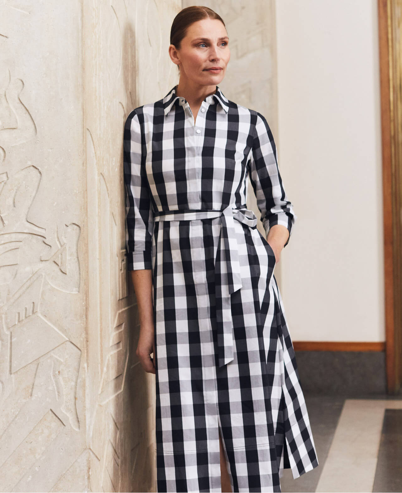 Hobbs stands next to art deco relief wearing a gingham print shirt dress.