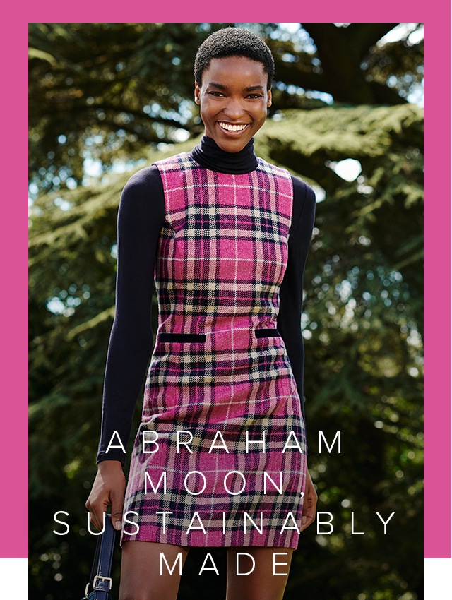 Alt text: Hobbs wool shift dress with a check pattern in pink shades made from Abraham Moon’s British made sustainable wool fabric, layered over a navy blue turtleneck women’s jumper styled with a leather tote bag.