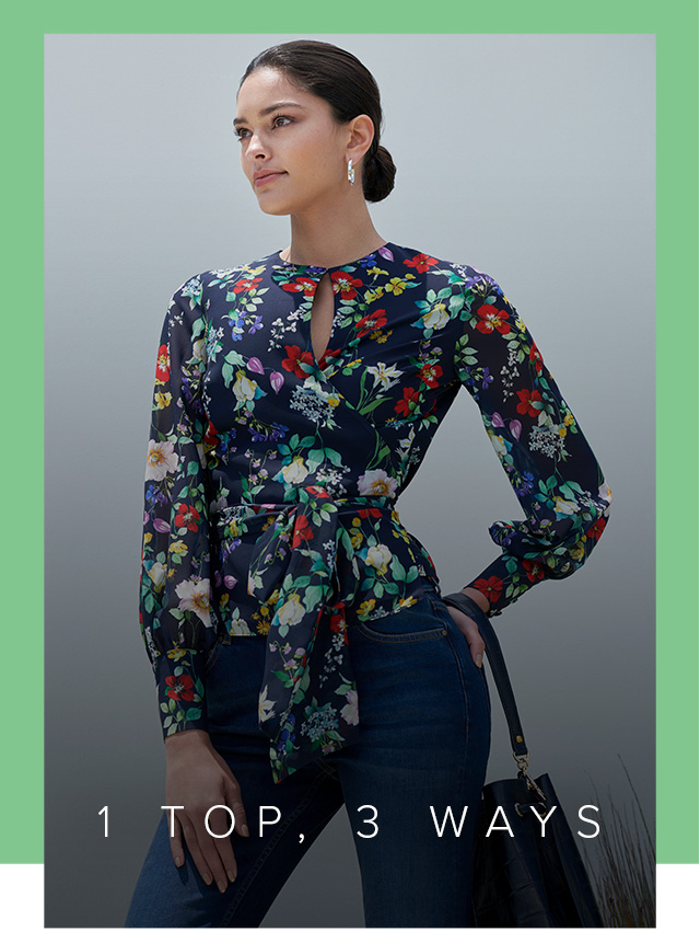 A brown haired woman poses in the Hobbs Meadow Blouse, a navy floral printed wrap top styled with denim jeans and a leather bucket bag