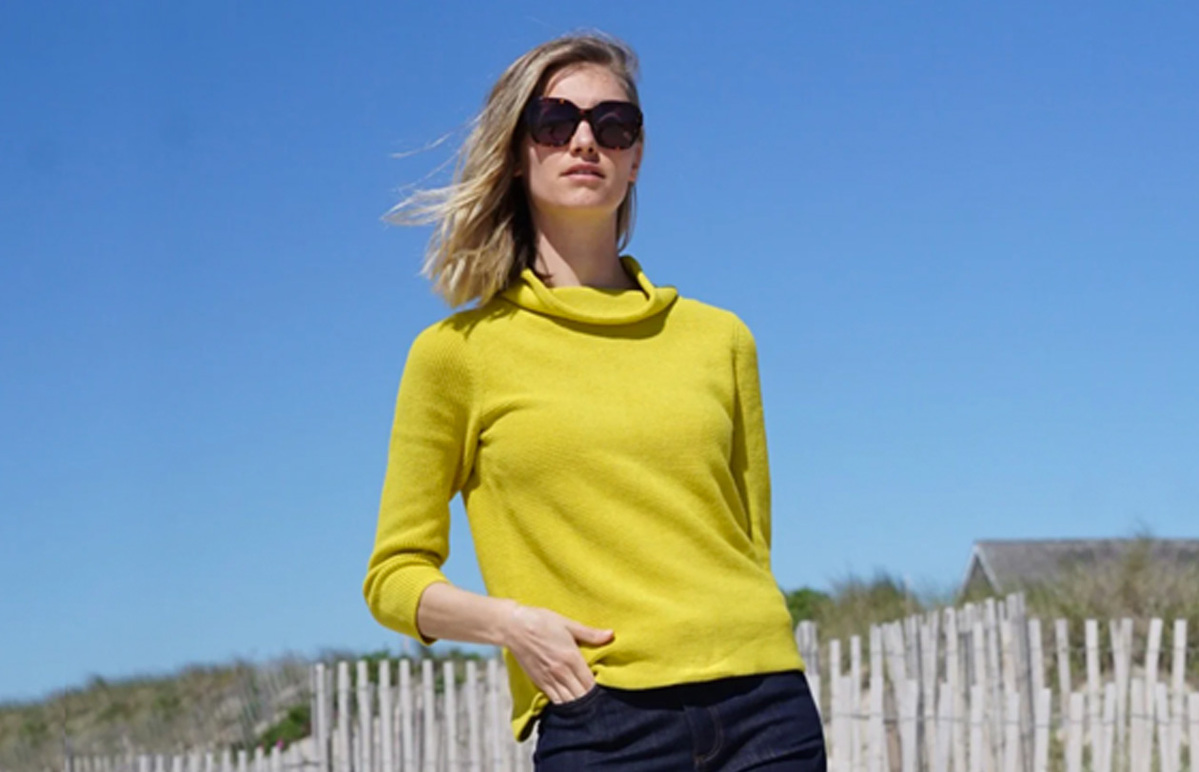 A woman poses in sunglasses, a bright yellow jumper wth dark denim jeans