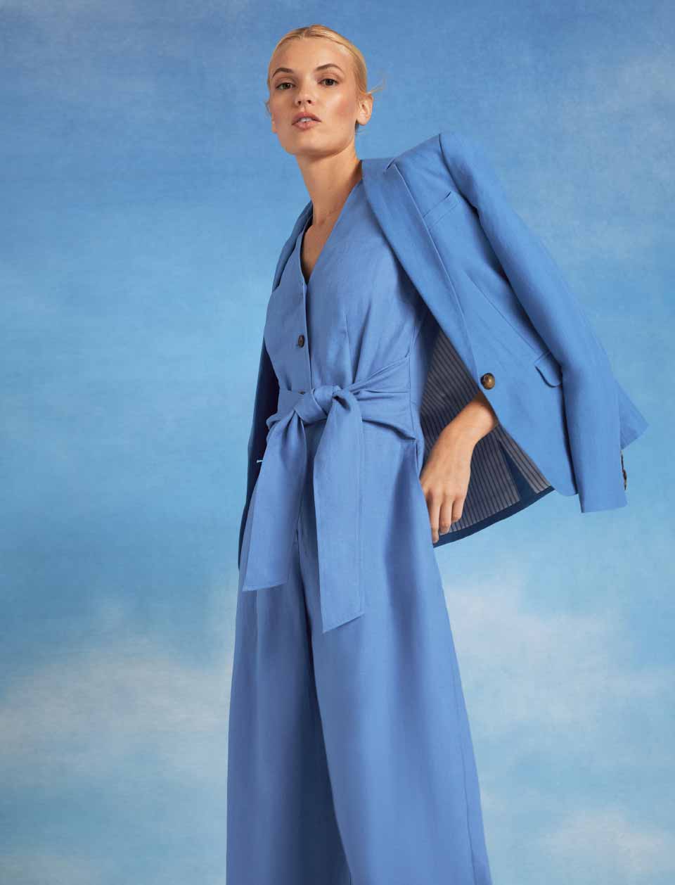 Close-up image of model pictured in front of a sky blue background wearing a blazer over a jumpsuit.