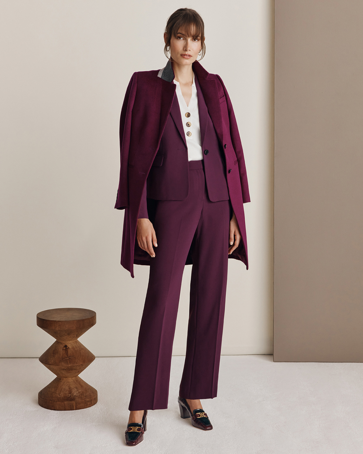 Model photographed against a neutral backdrop wearing a Hobbs burgundy suit with matching coat draped on her shoulders.