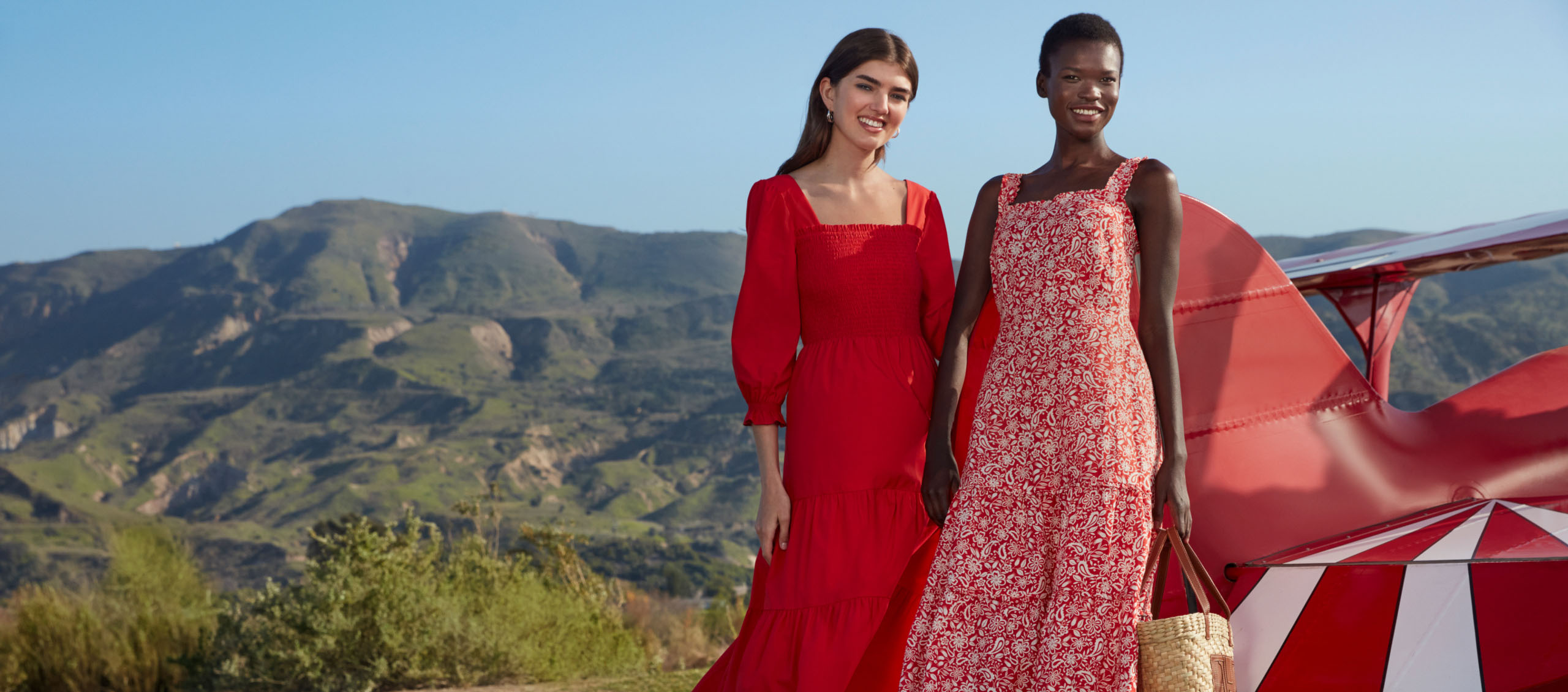 Image of two models pictured in front of an aeroplane wearing open-toe sandals, summer dresses and woven tote handbags.