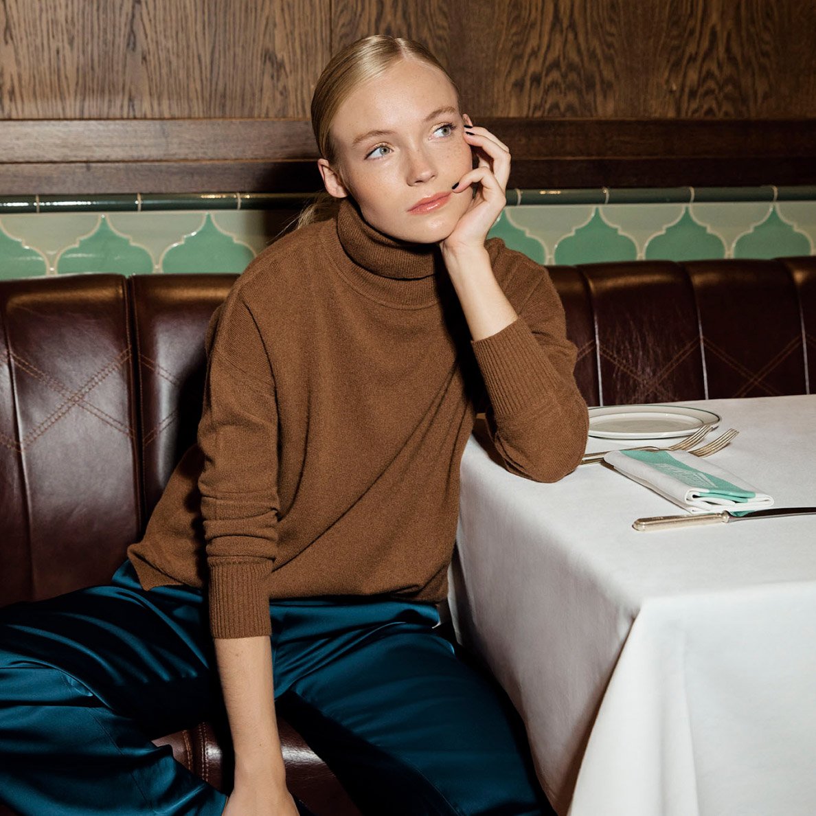 Model photographed leaning against a sofa and wearing a blouse top, trousers and sandals