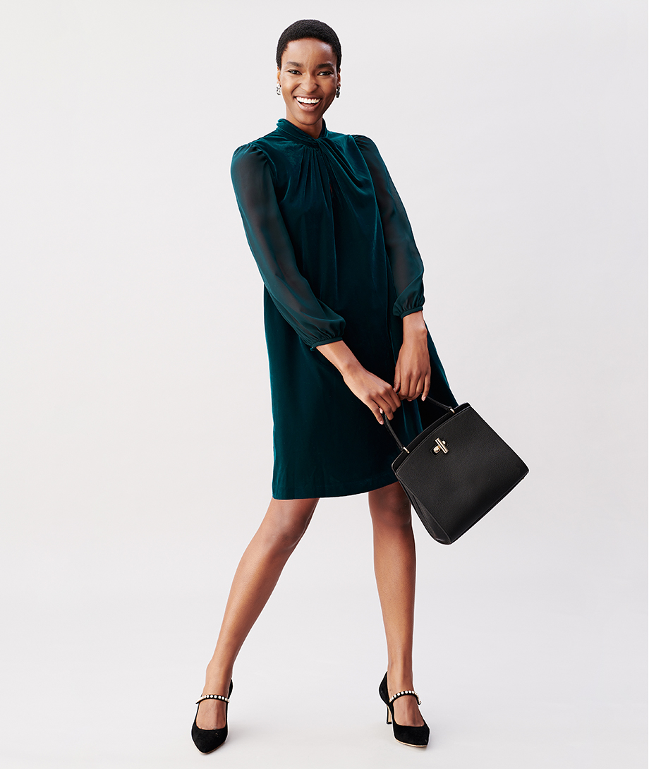 Hobbs model wearing a green velvet dress, paired with black heels with an embellished strap design and a black leather bag.