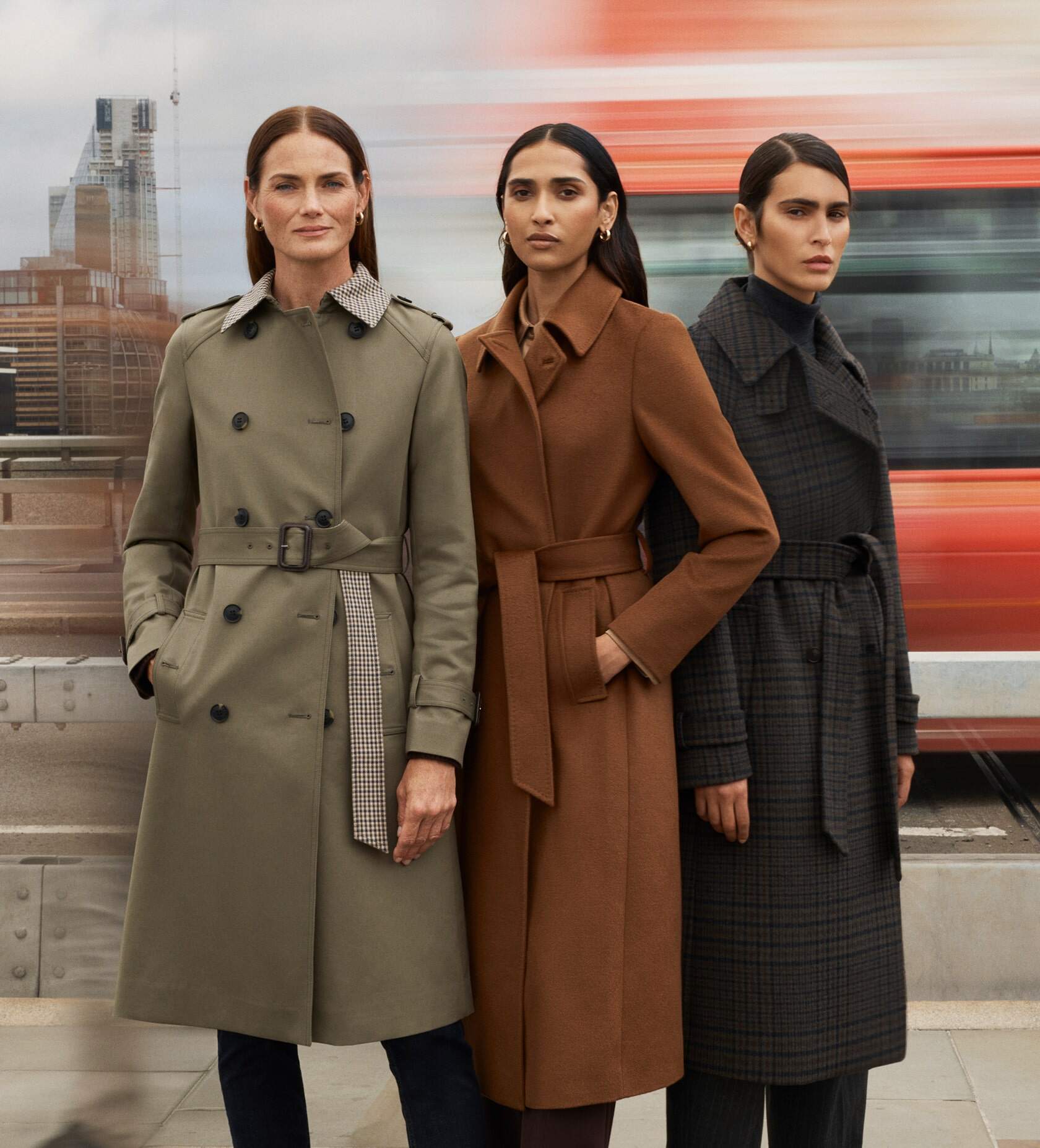 Three models wearing coats stand on a busy street as a bus speeds past in the background.