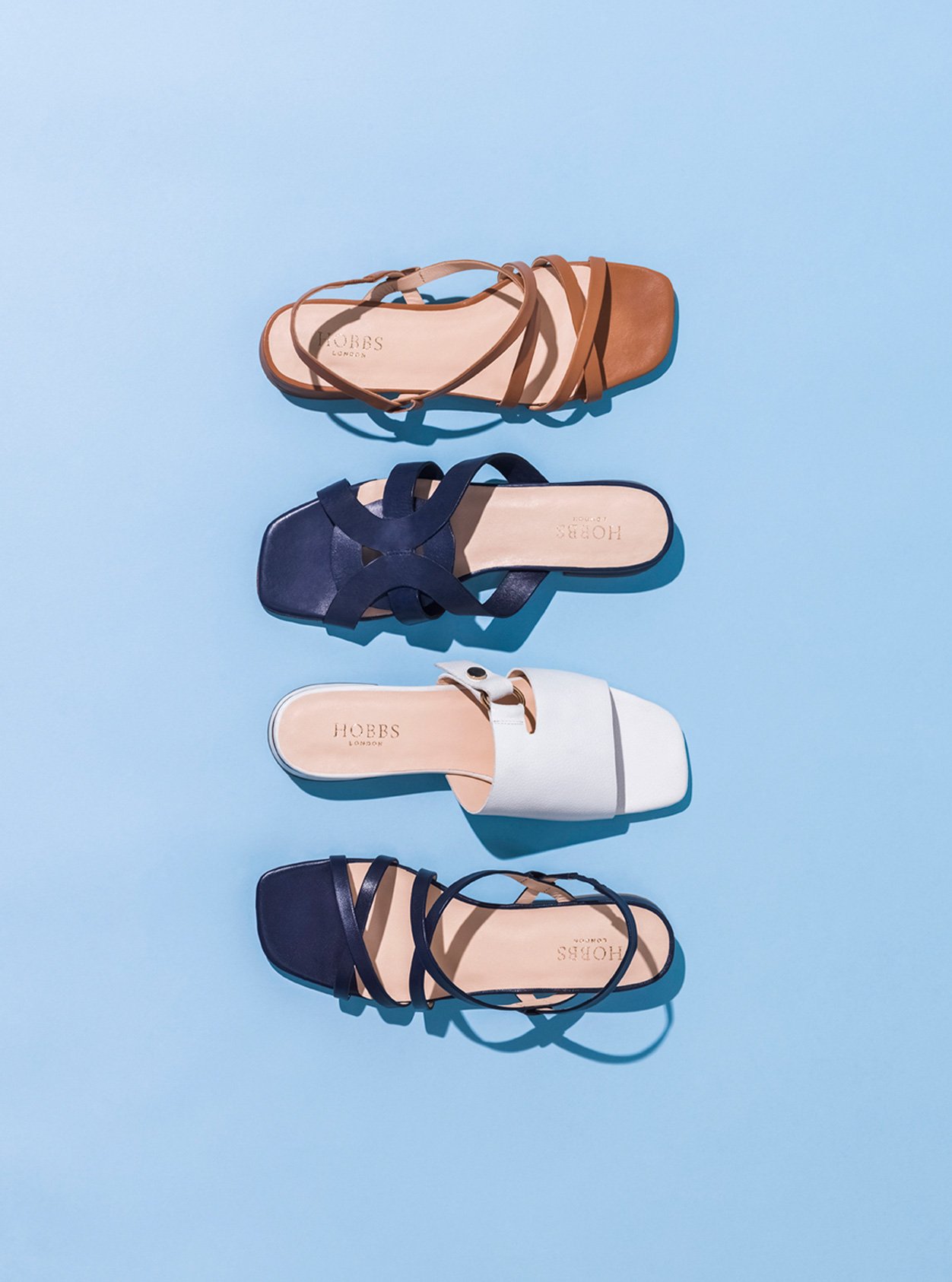 Four summer sandals from Hobbs against a blue background, from top to bottom: Brown sandal, dark blue sandal, white sandal, black sandal.