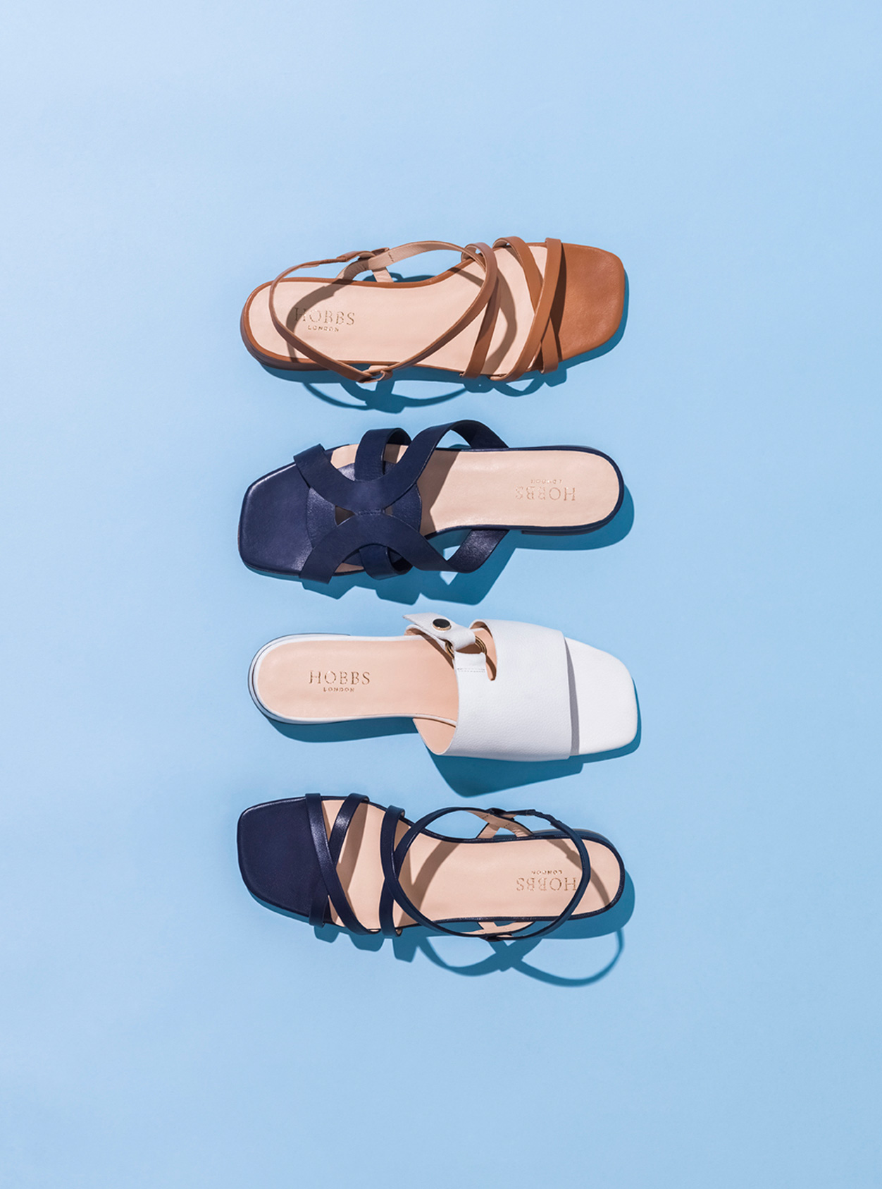 Four summer sandals from Hobbs against a blue background, from top to bottom: Brown sandal, dark blue sandal, white sandal, black sandal.