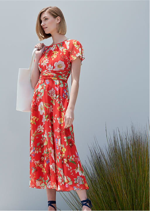 Woman stands in a red floral midi dress wearing navy blue espadrilles and holding a white leather tote bag