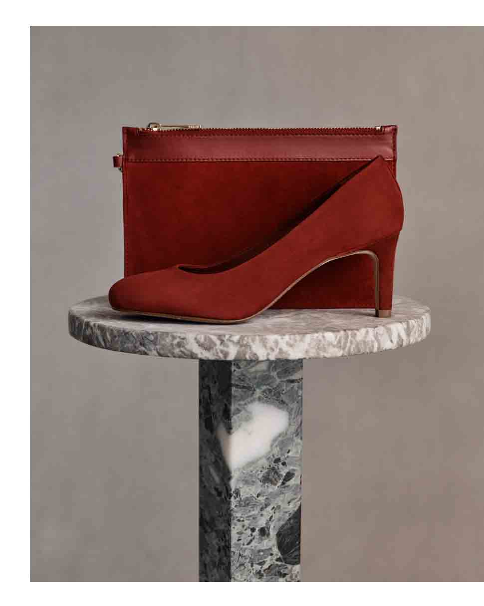 A burgundy suede court shoe and clutch bag sit atop a marble plinth