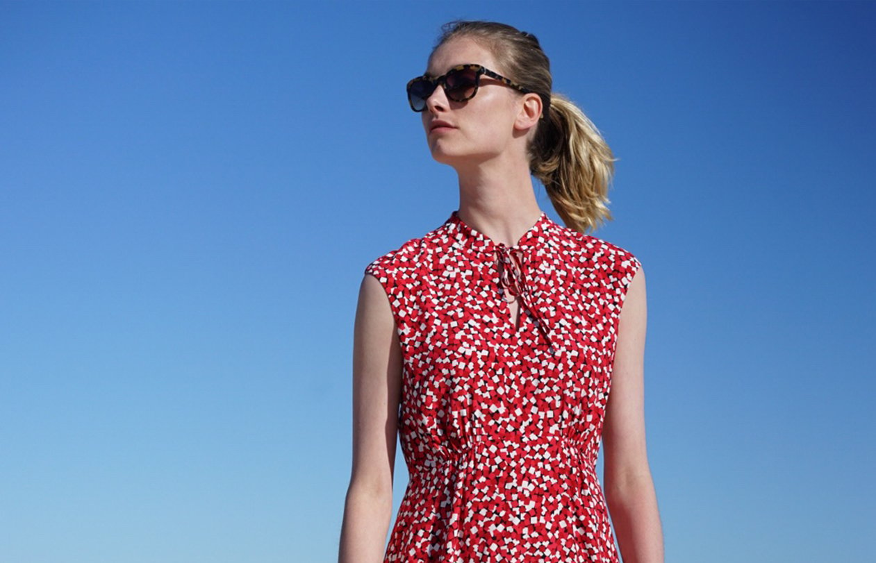 A Hobbs model wears a red dress with short cap sleeves and a neck fastening at the front, styled with sunglasses with blue sky in the background.