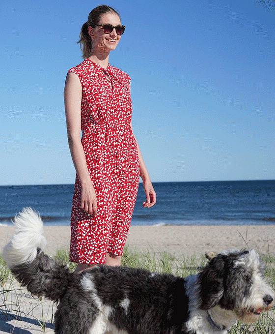 Hobbs model, Irish Van Berne,photographed on the beach in a red floral sleeveless dress with a grey and white dog. 