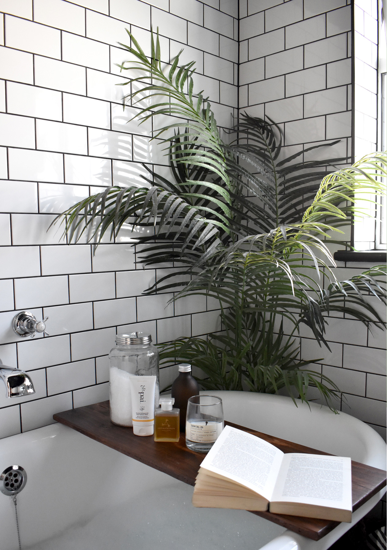 A serene white bathroom with white metro tiles and black grout, a green leafy plan and an array of luxurious bath products.