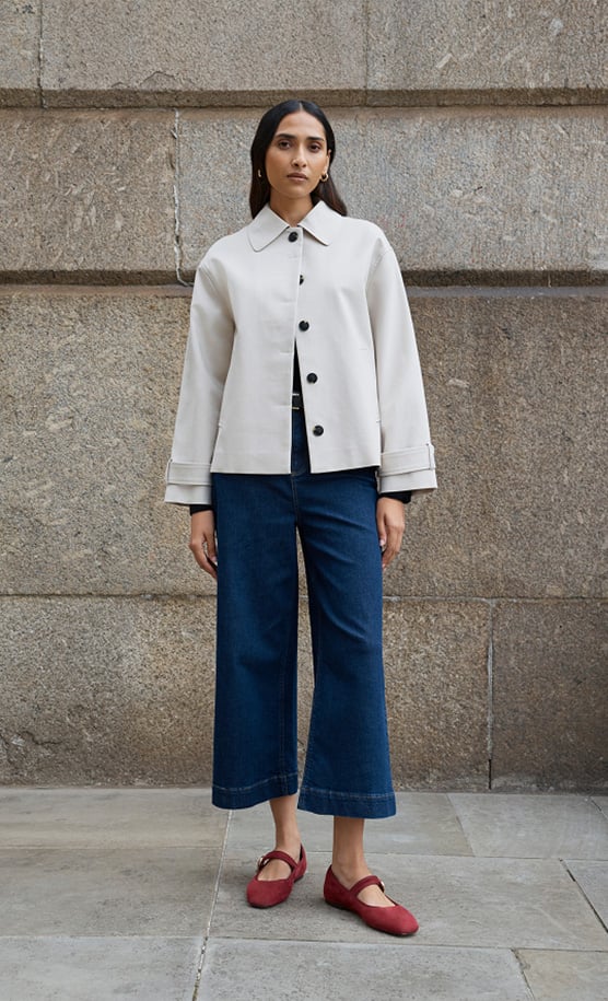 Model stands against a wall wearing a grey cropped trench.