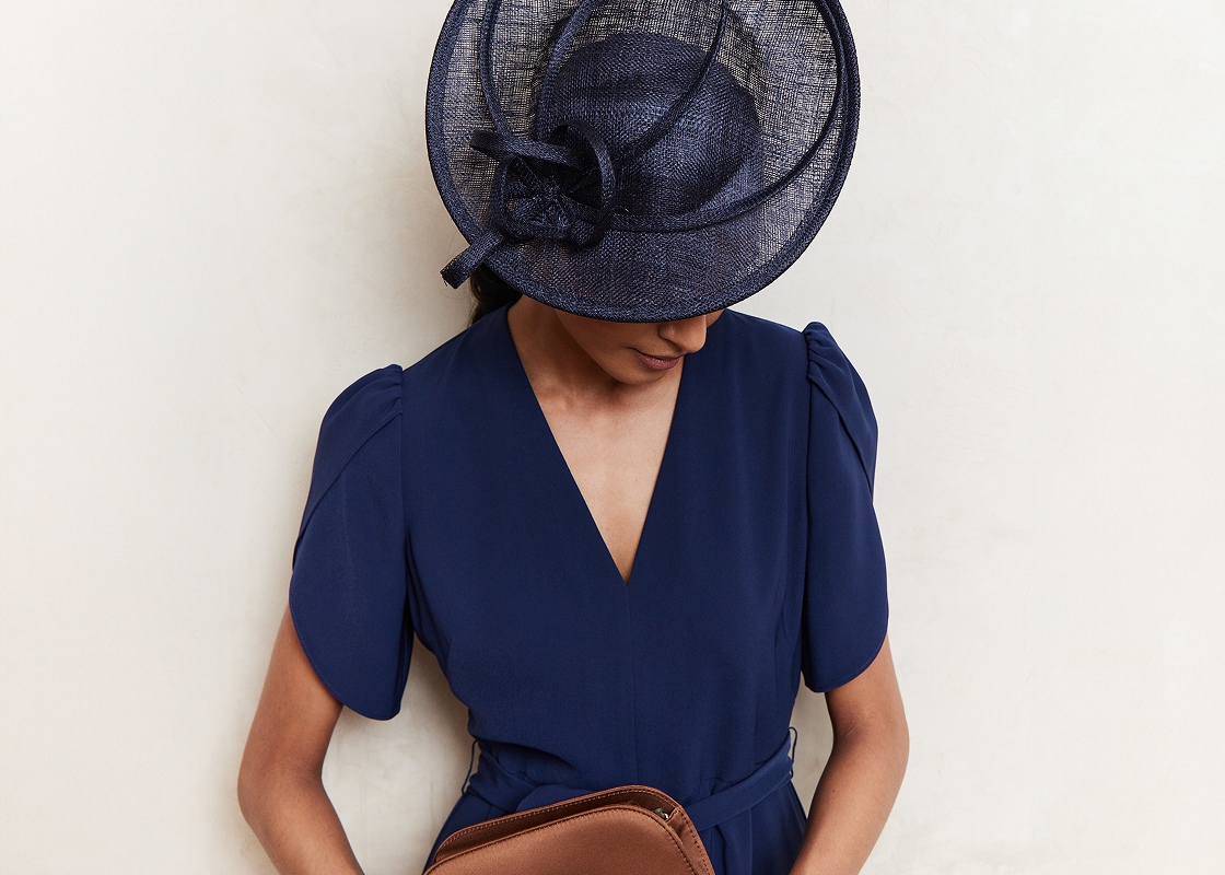 An image of a model wearing a navy occasion dress and coordinating fascinator.