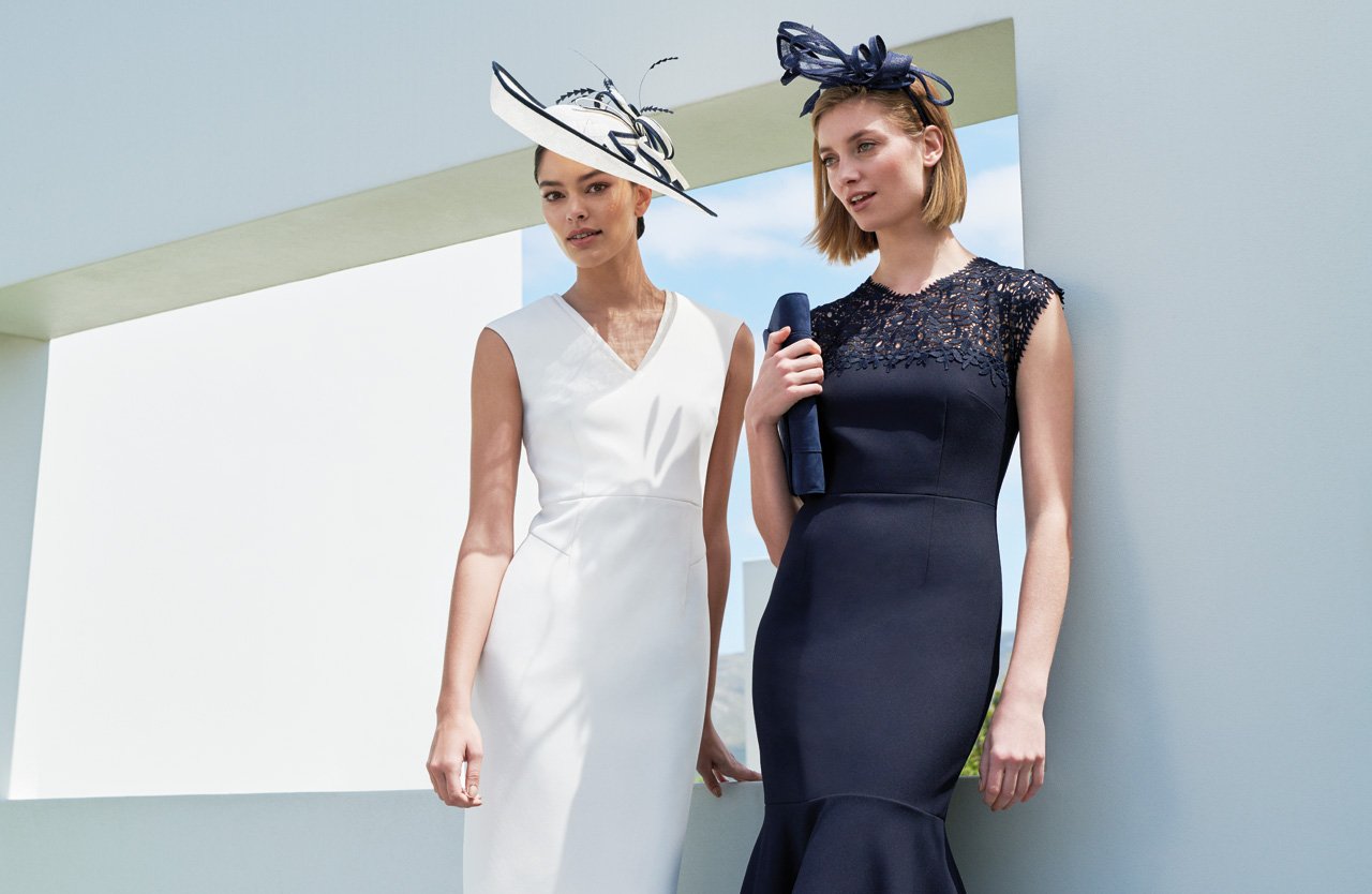 Two Women Pose in Ivory and Navy Event Dresses Wearing Fascinators