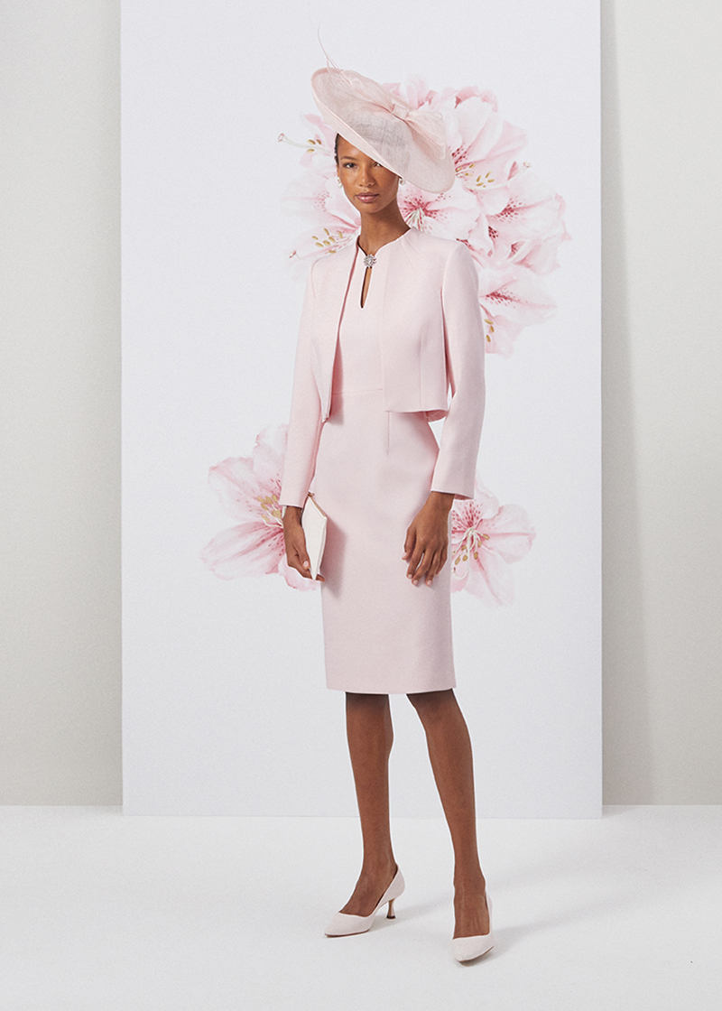 Image of model standing in front of a floral painted background wearing a matching pale pink jacket and dress two piece suit.