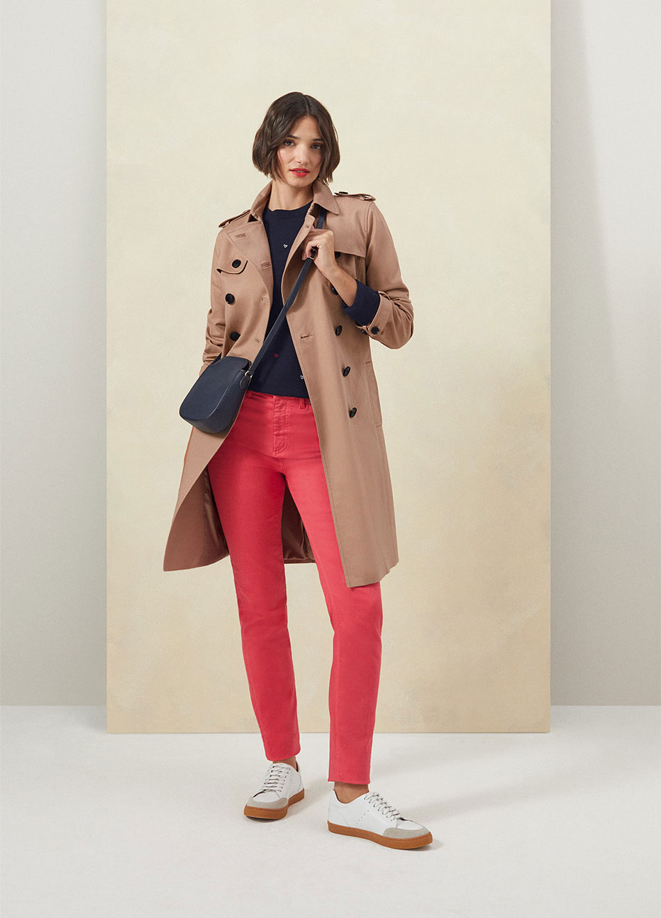 Model photographed against a yellow backdrop wearing a Hobbs trench coat, navy jumper and coral pink jeans.