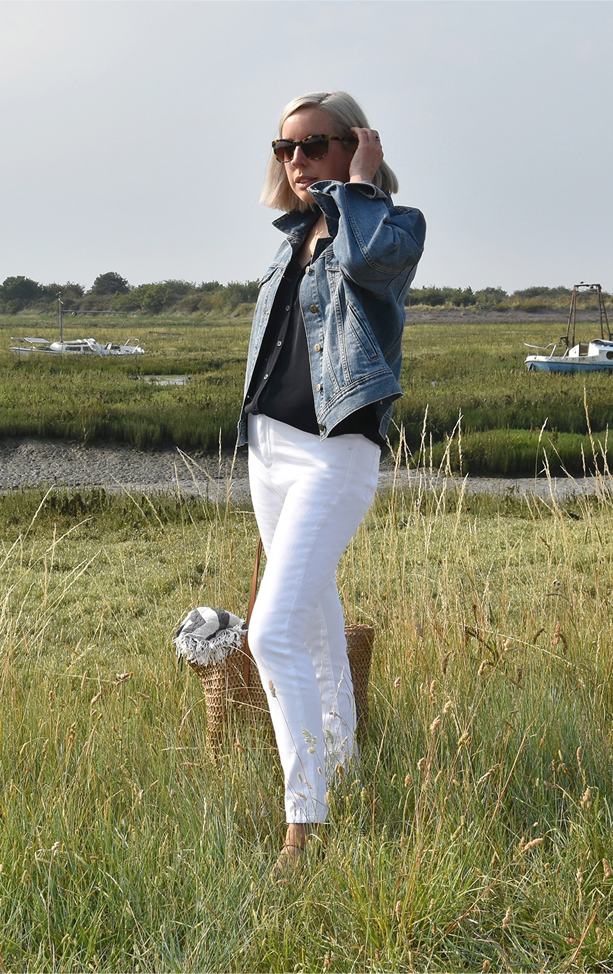 Jen Wilkins, Creative Copy & Editorial Manager at Hobbs stands in a secluded spot by the coast wearing a blue denim jacket, a navy blue blouse, white denim jeans and tan sandals styled with a straw bag.