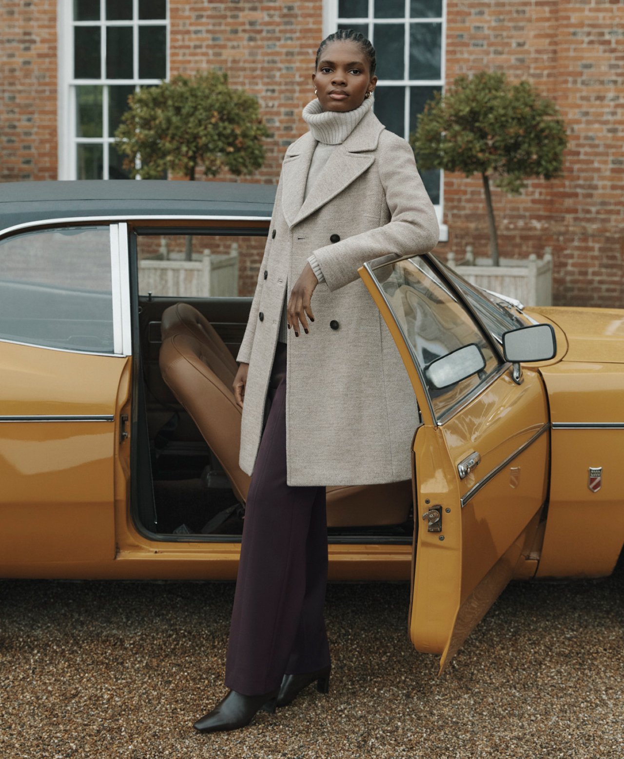 A woman wearing a beige coat layered over a high neck jumper and brown trousers.