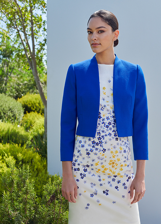 Woman poses in white formal shift dress witha blue and yellow floral pattern with a matching cobalt blue jacket