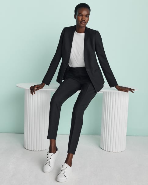 Hobbs model leans on a white column table wearing a black trouser suit, white t-shirt and trainers.