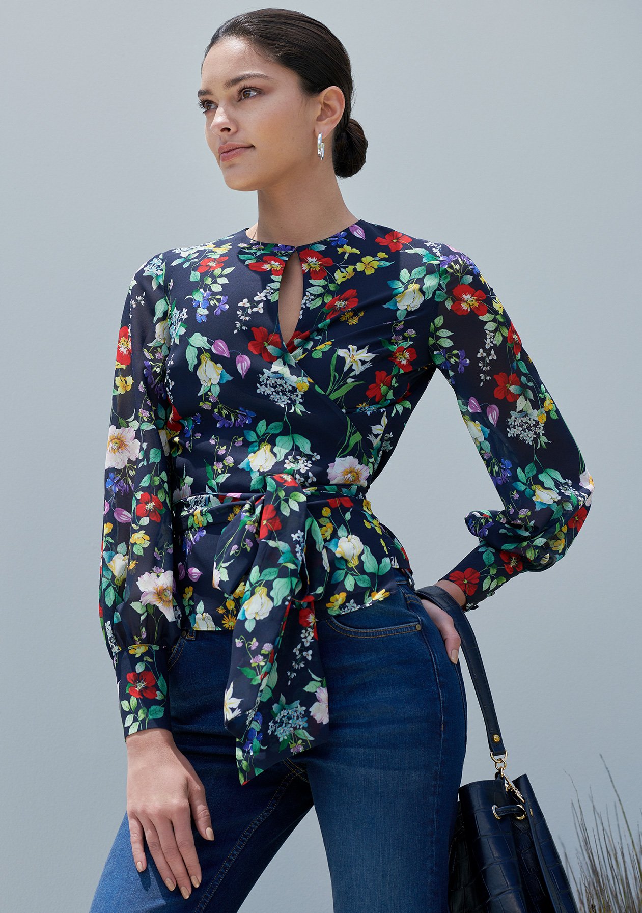 A brown haired woman poses in the Hobbs Meadow Blouse, a navy floral printed wrap top styled with denim jeans and a leather bucket bag