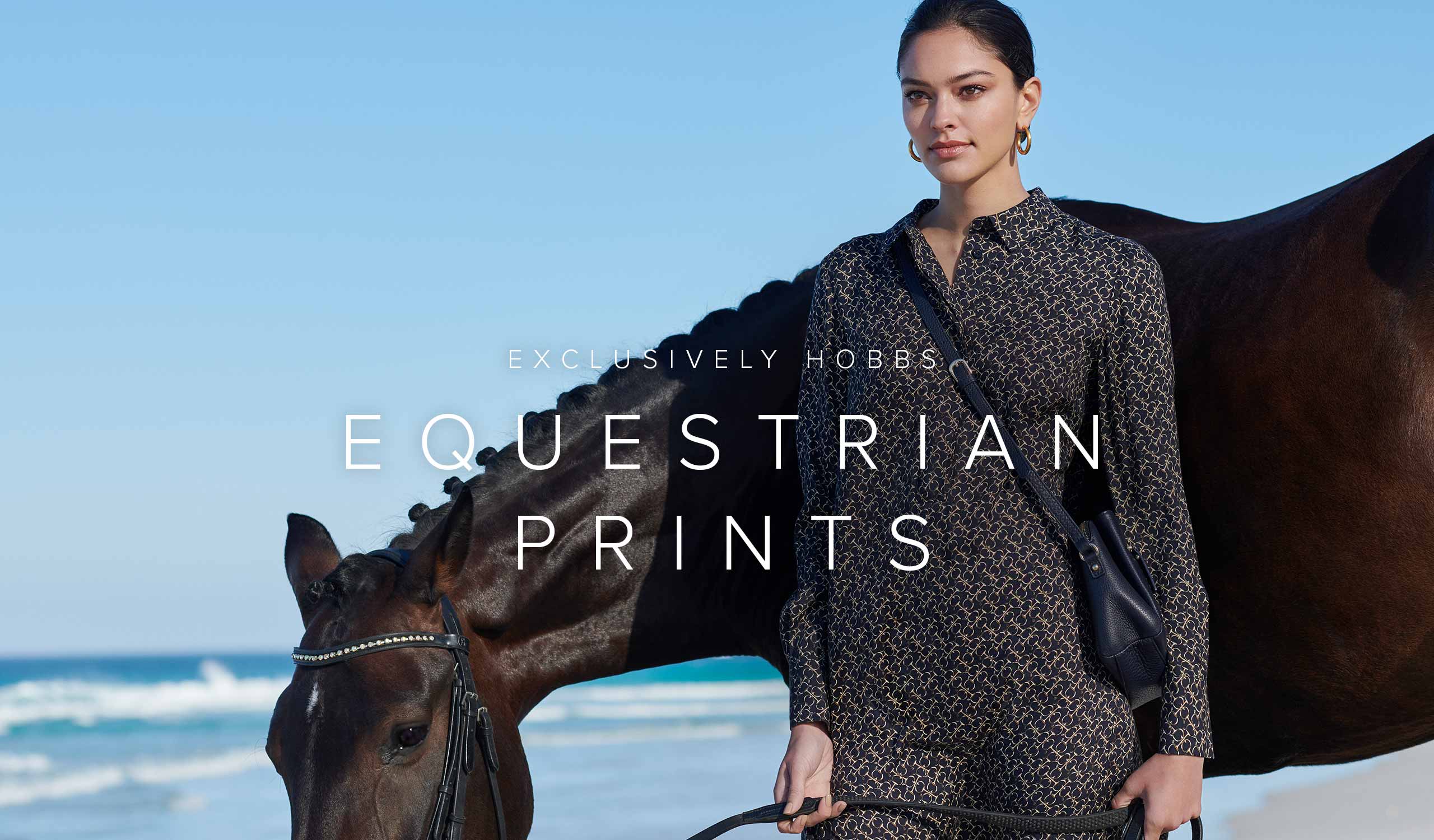 Woman poses in an  equestrian print, green and navy blue shirt dress with a black horse on a beach.