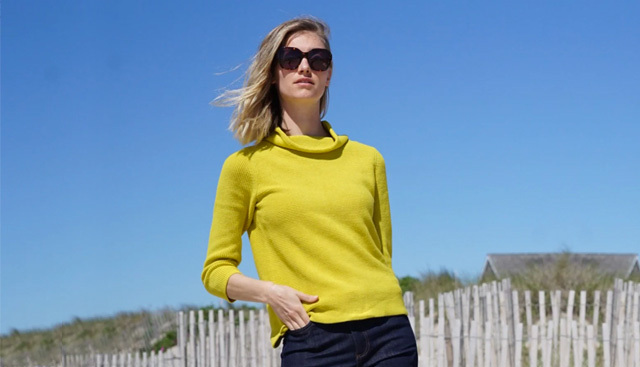 A woman poses in sunglasses, a bright yellow jumper wth dark denim jeans