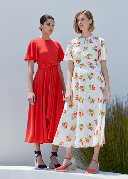 Two woman poses, one wears a flame red midi dress with sleeves and the other a white midi dress wth a red and yellow floral motif with red espadrille sandals