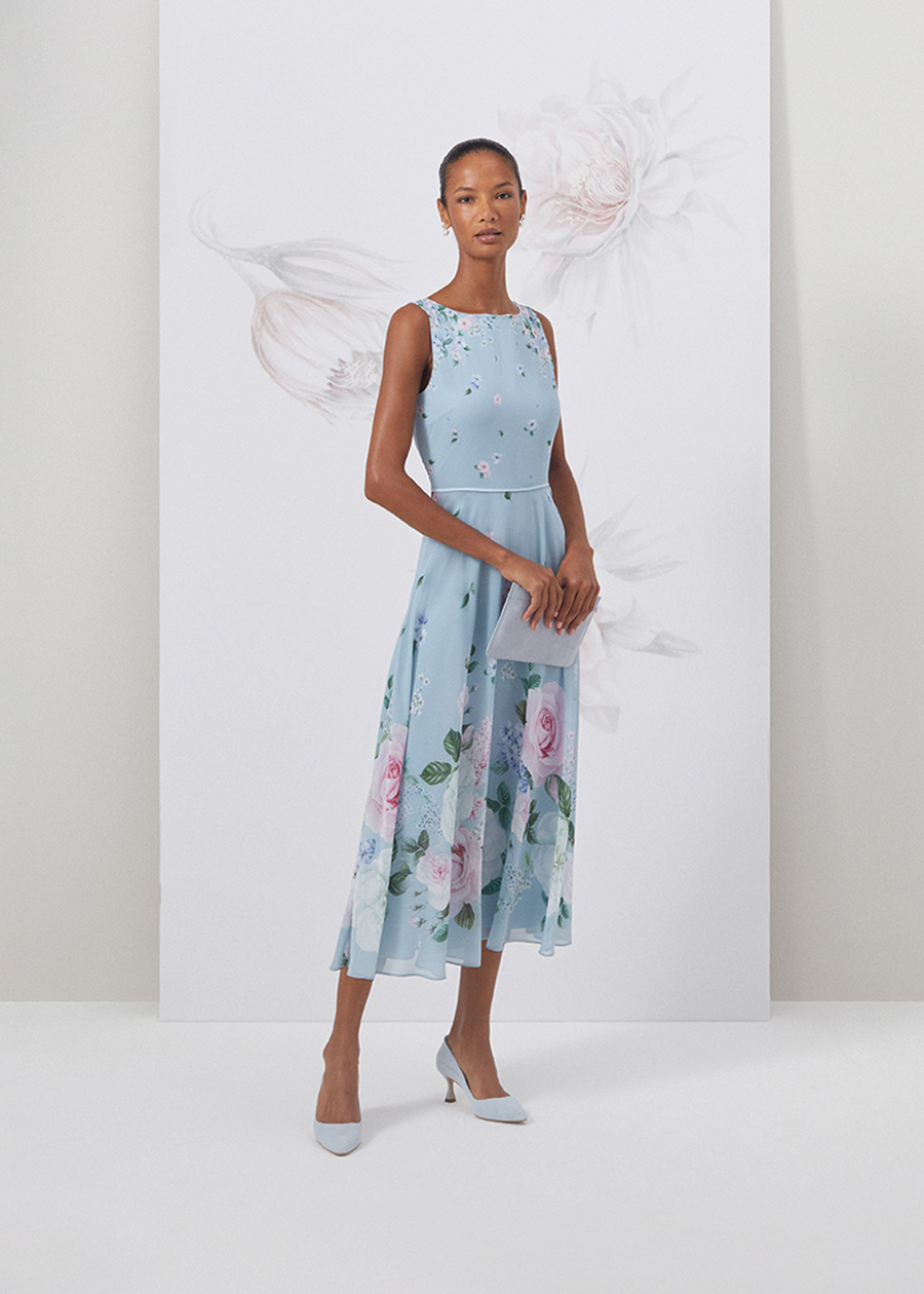 Close up image of model standing in front of a floral painted background wearing a navy floral print dress with matching navy fascinator.