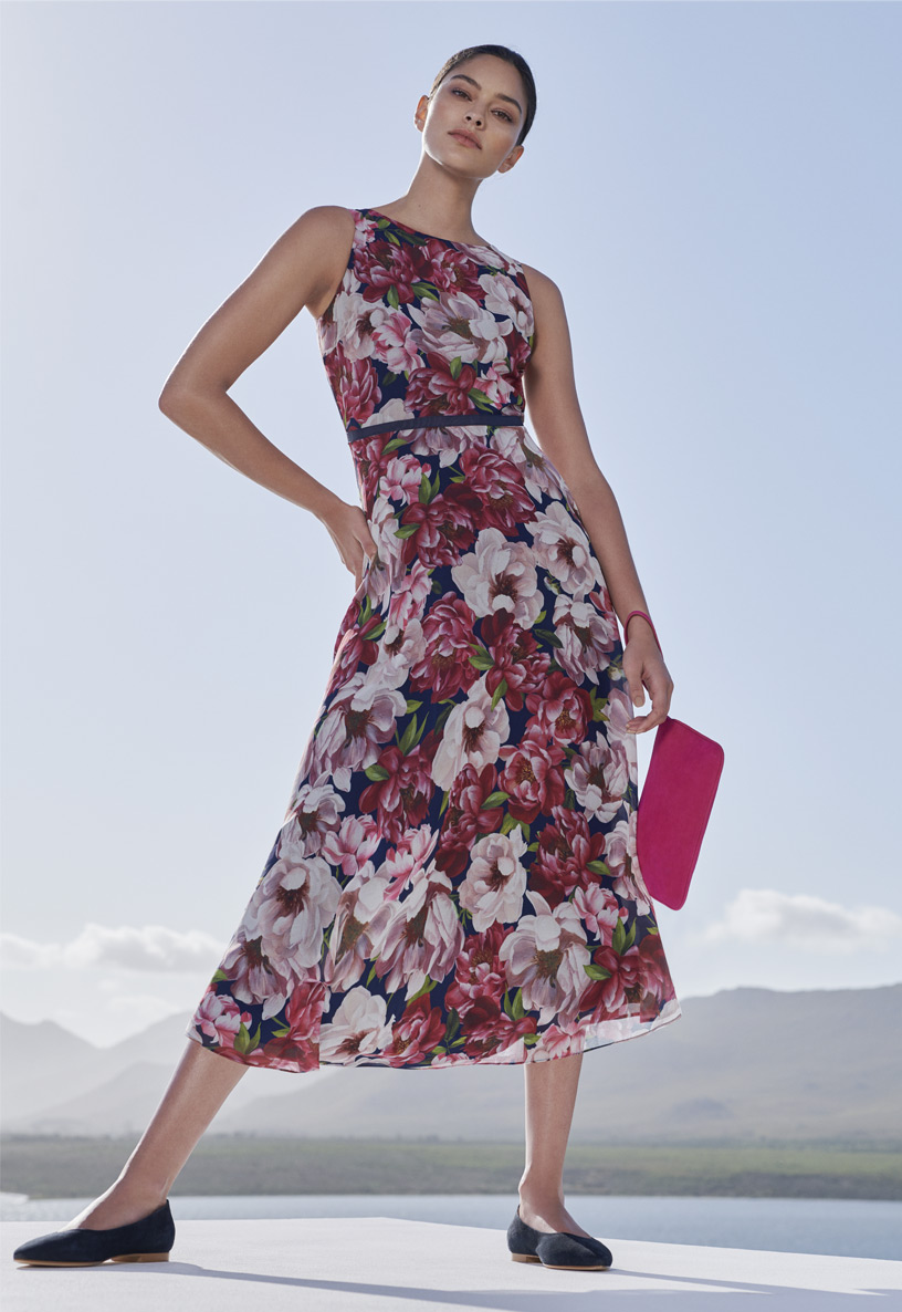 Woman poses in floral midi dress with a bright pink suede clutch bag and flat navy blue suede smart shoes.