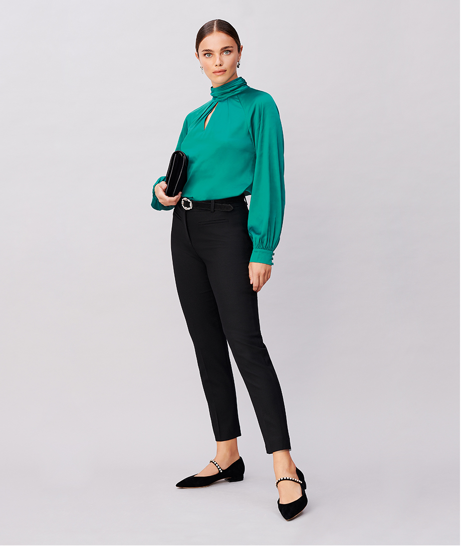 Hobbs model wearing a roll neck satin blouse with cuff sleeves in green, styled with a black belt with an embellished buckle, black slim trousers for women, a black clutch and black pointed flat shoes with a embellished strap design.