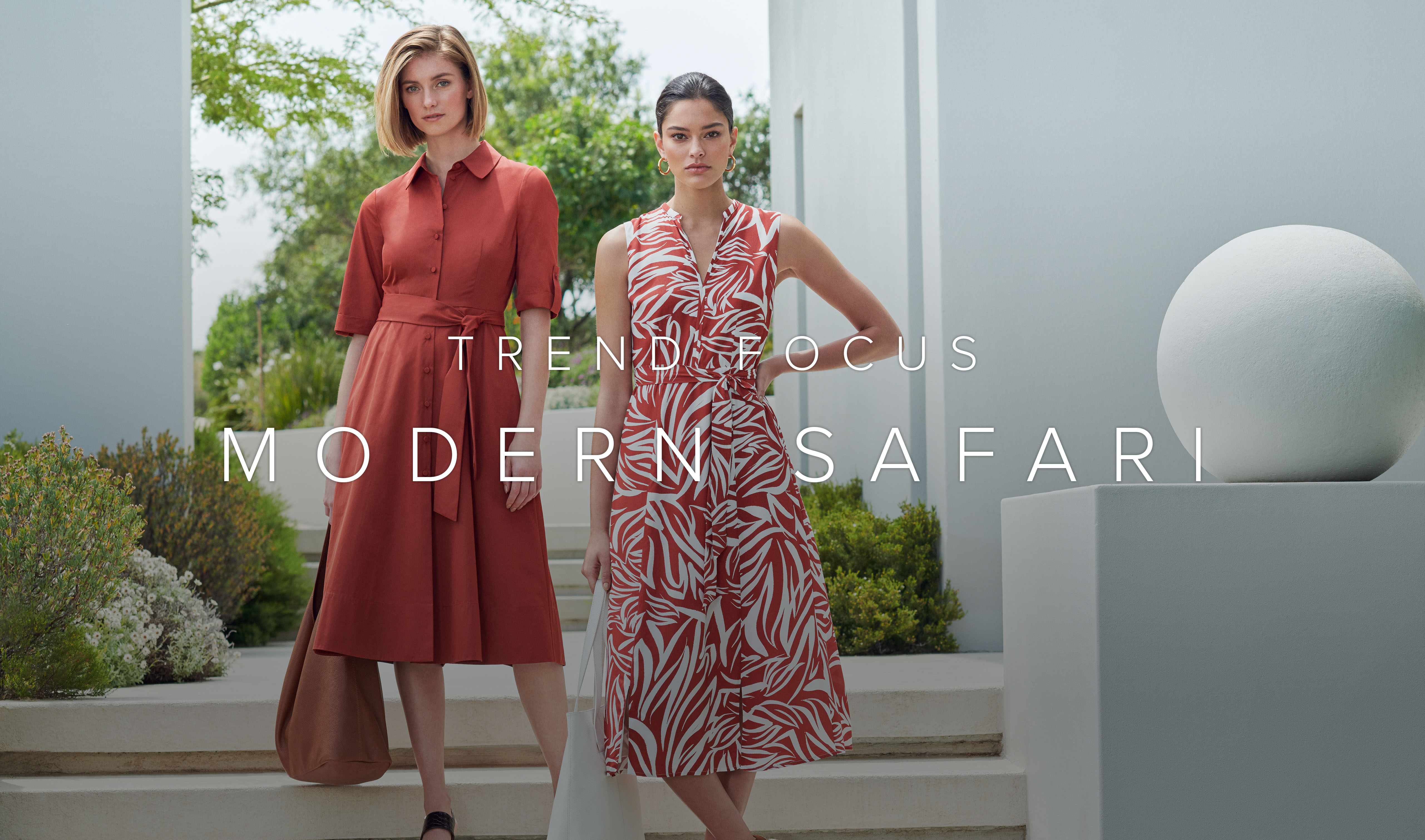 Two women pose against a modernist architectural background. One wears a brown knee-length shirt dress and the other a rust brown and wite patterned midi length dress.