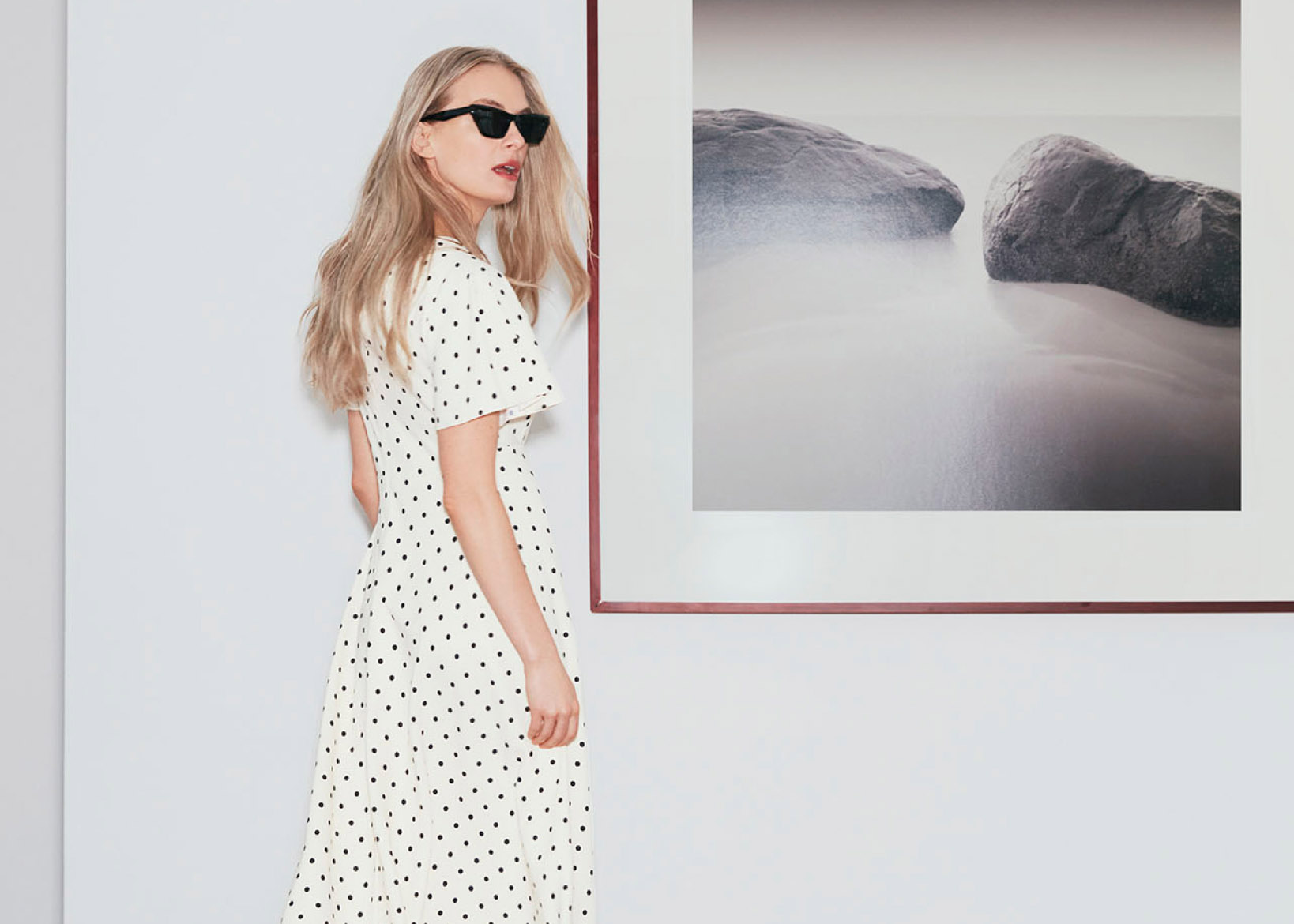 Model photographed standing in front of a photo on a wall wearing a polka dot dress.