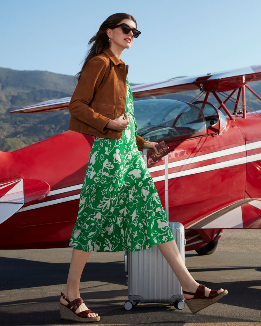 Image of a model pictured in front of an aeroplane and wearing a floral summer dress and wedge sandals.