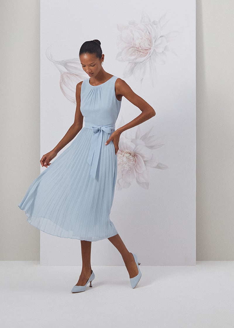 Image of model standing in front of a floral painted background wearing a pale blue pleated midi length dress.