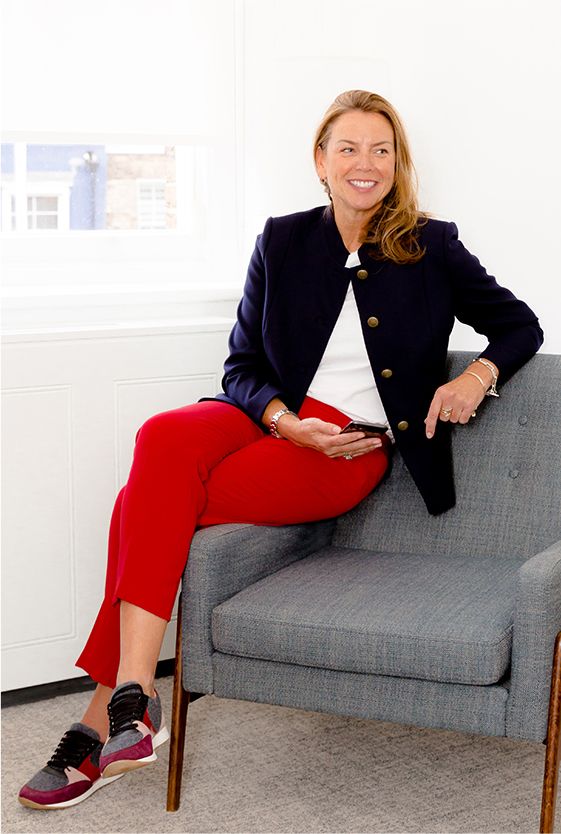 Hobbs' Product Director, Sally Ambrose, photographed sitting on a chair wearing a jacket, trousers and trainers.