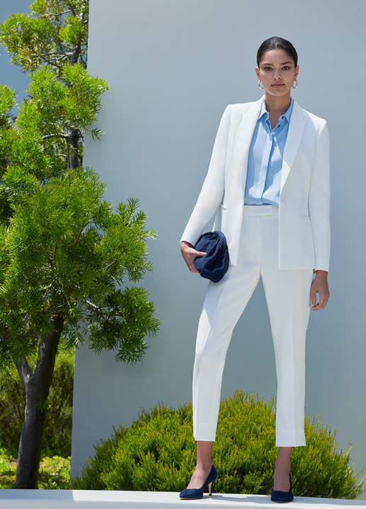 Woman stands in a white trouser suit styled with a pale blue shirt and navy suede court shoes and matching clutch bag