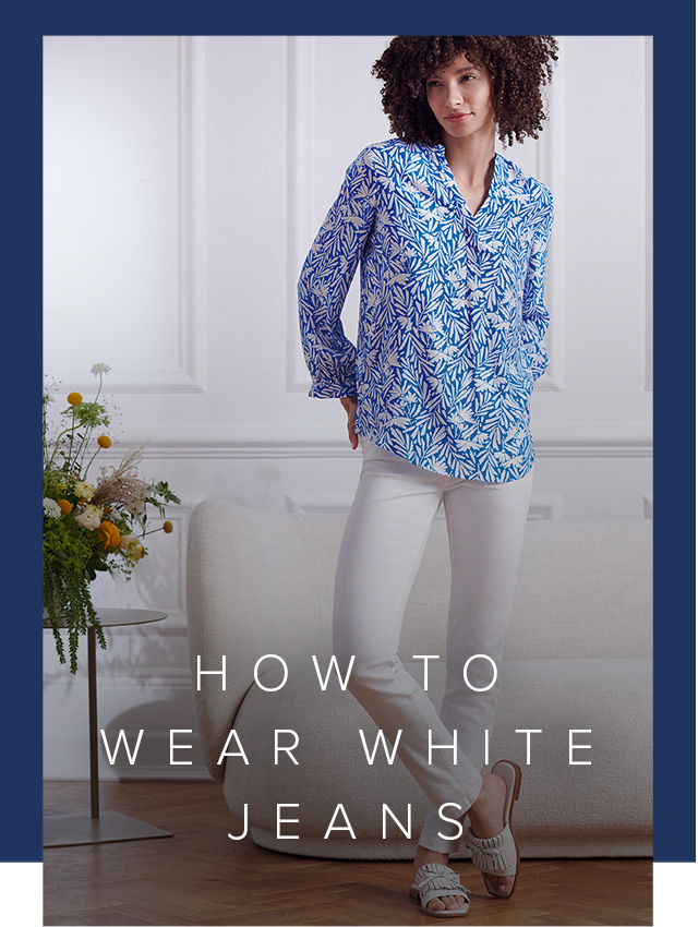 Model wearing white jeans and a blue printed blouse stands next to a sofa and vase of flowers.