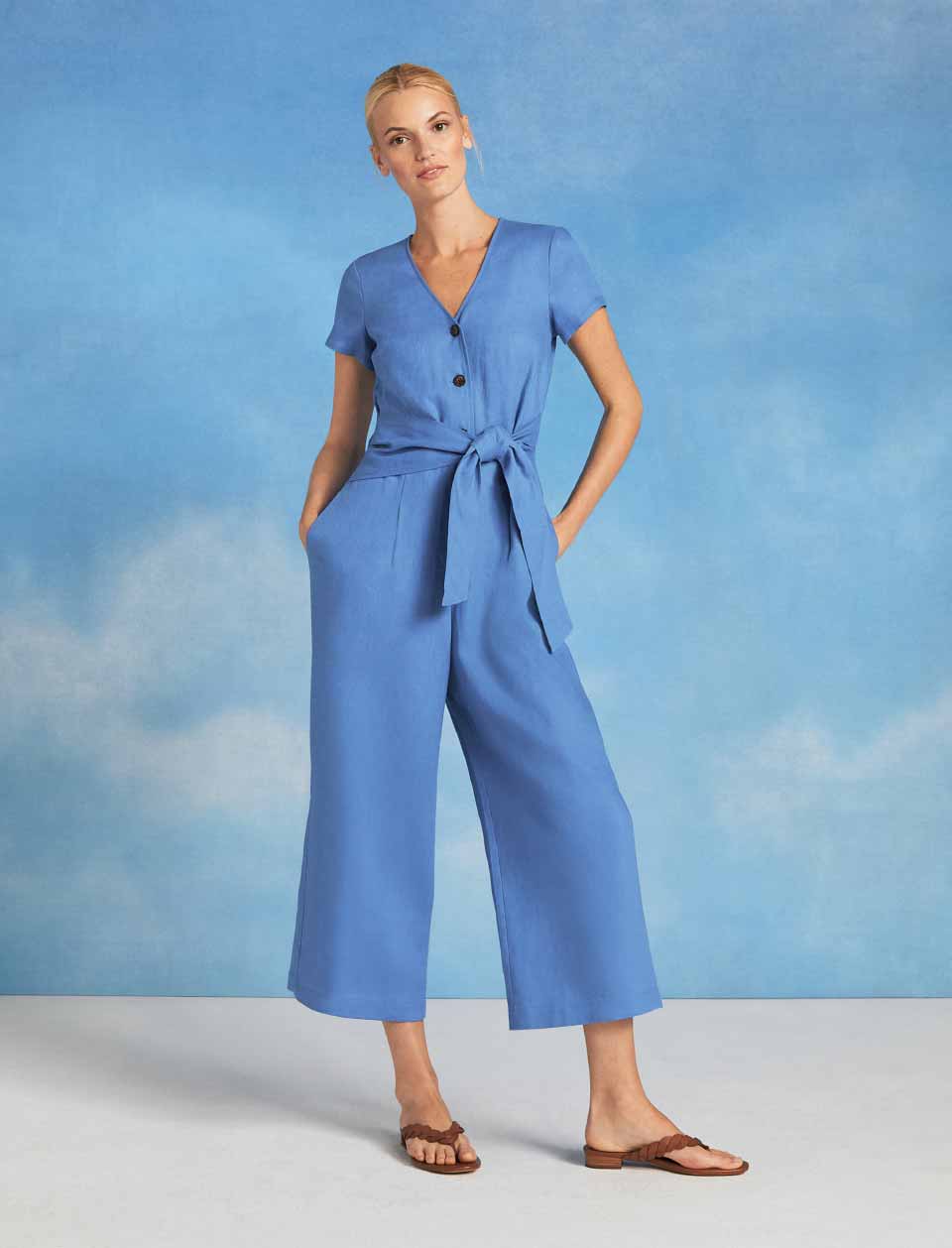Model photographed in front of a sky blue background wearing a linen jumpsuit.