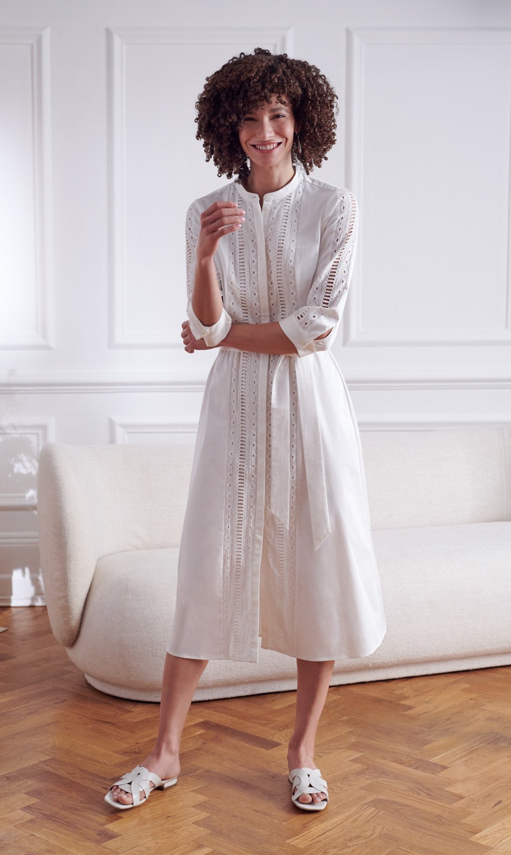 Model wearing a white cotton midi dress and leather slides stands beside a white boucle sofa.