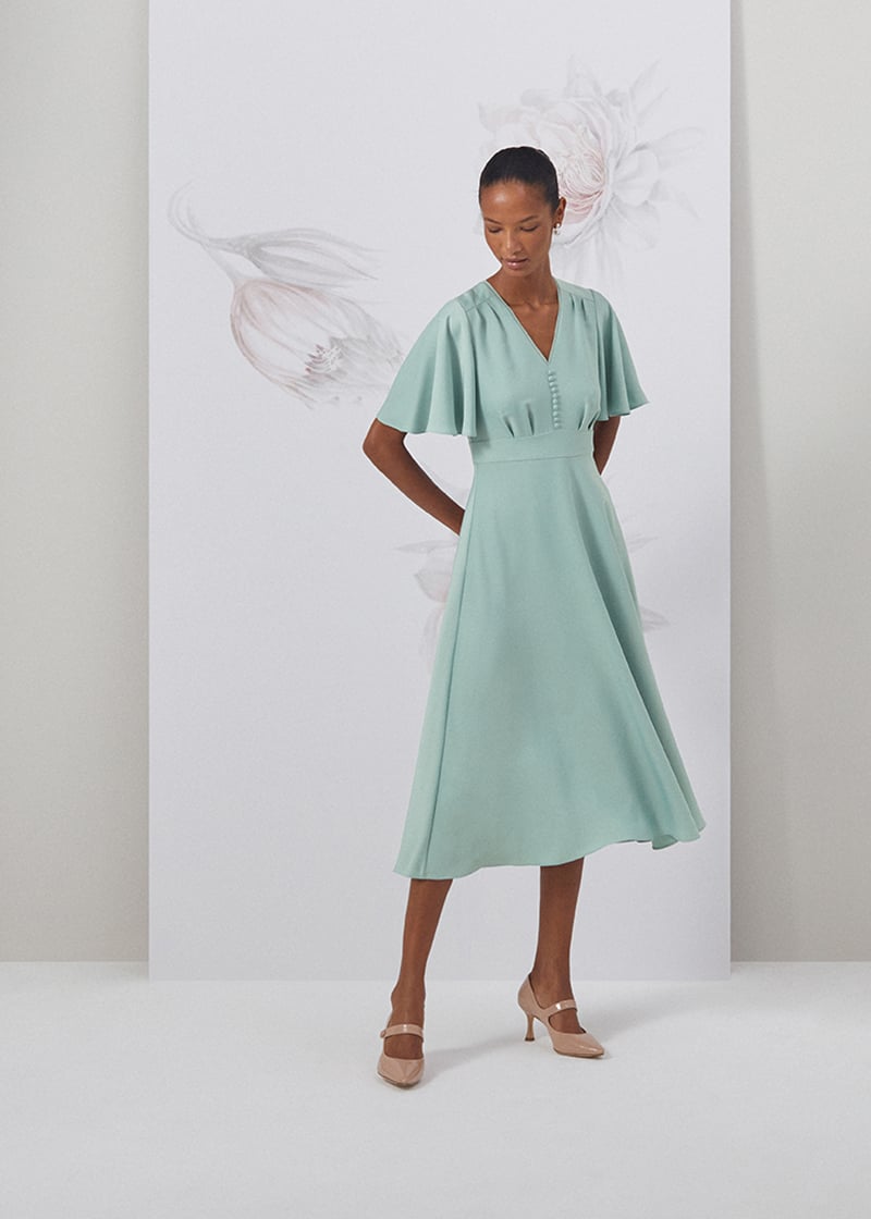 Image of model standing in front of a floral painted background wearing a sage green fit and flare midi length dress.